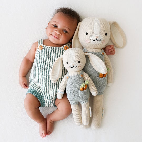 White background with baby laying, holding both sizes of Henry The Bunny by Cuddle and Kind