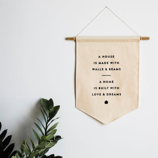 White background with a sprig of greenery in the bottom left hand corner and a House and Home Canvas Banner by Gladfolk. Banner is black font that says "A house is made with Walls & Beams ---- A Home is build with love & dreams"