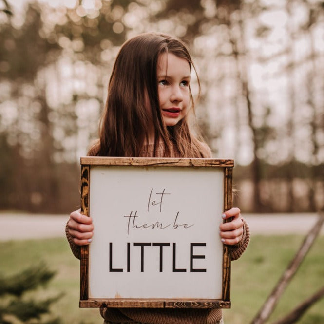 Little girl standing in grass in front of trees with the Let Them Be Little Wood Sign by Restored Signs & Decor. Sign is white and says "Let them be little" in black, with a dark wood frame.