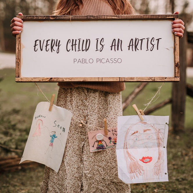 Little girl standing outside, grass and wood behind her, she's holding the "Every Child Is An Artist Wood Sign by Restored Signs & Decor".