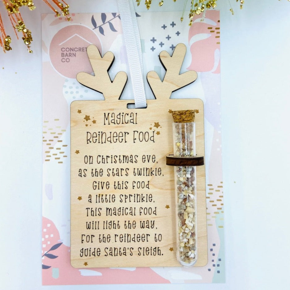 White background with the Reindeer Food Ornament by Concrete Barn in its packaging. Ornament is square, with reindeer ears, engraved writing, and a small vial of "Reindeer Food".