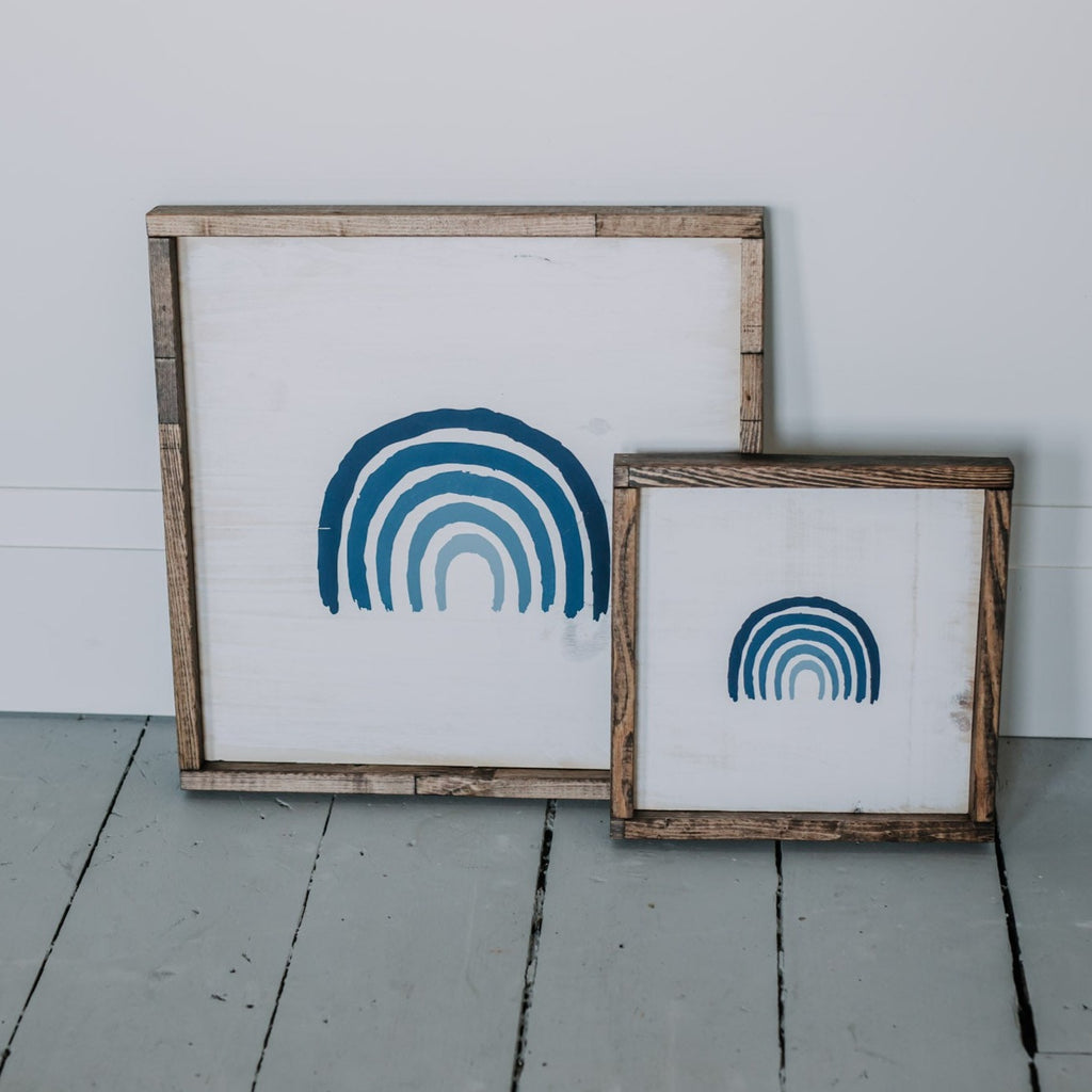 White wall with a grey painted floor, and the Rainbow: The Tennyson Wood Sign by Restored Signs & Decor. Sign is white with a blue gradient rainbow and a wood frame.