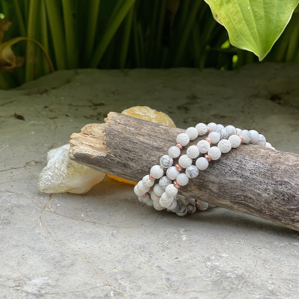 Cement with greenery in the back, and a wood log with 3 White Howlite Diffuser Bracelets by Earth & Ether Co. around it. Bracelets are made from white howlite, and have rose gold spacers.