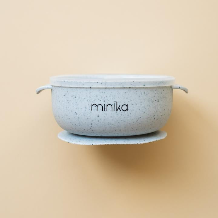 Beige background with a Silicone Bowl with Lid in Ice by Minika. Bowl is ice blue speckled, has a silicone base to stick to your table, and a clear lid.