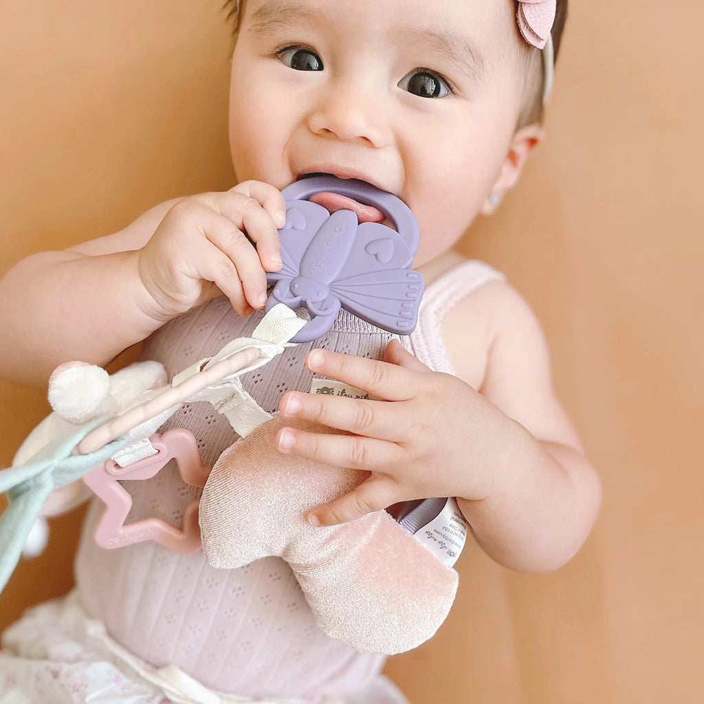 Beige/Brown background with baby girl laying, chewing on the Bitzy Busy Ring Teething Activity Toy Bunny by Itzy Ritzy.