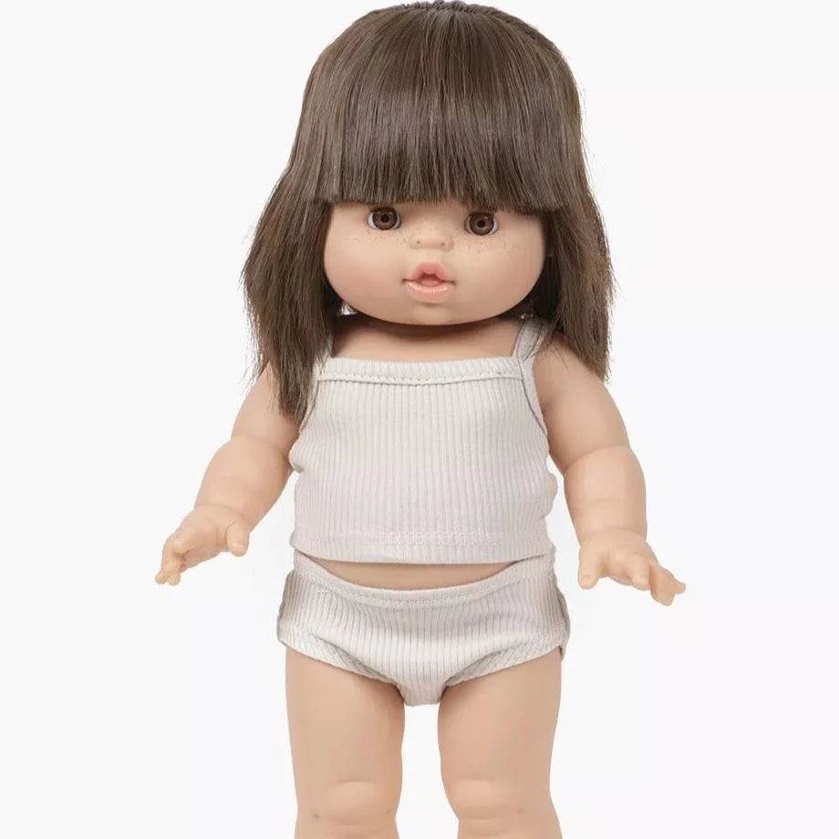 White background with Janelle Doll by Minikane. Doll has brown hair with bangs, light skin, brown eyes, and is wearing a linen outfit with a tank top & matching underwear.