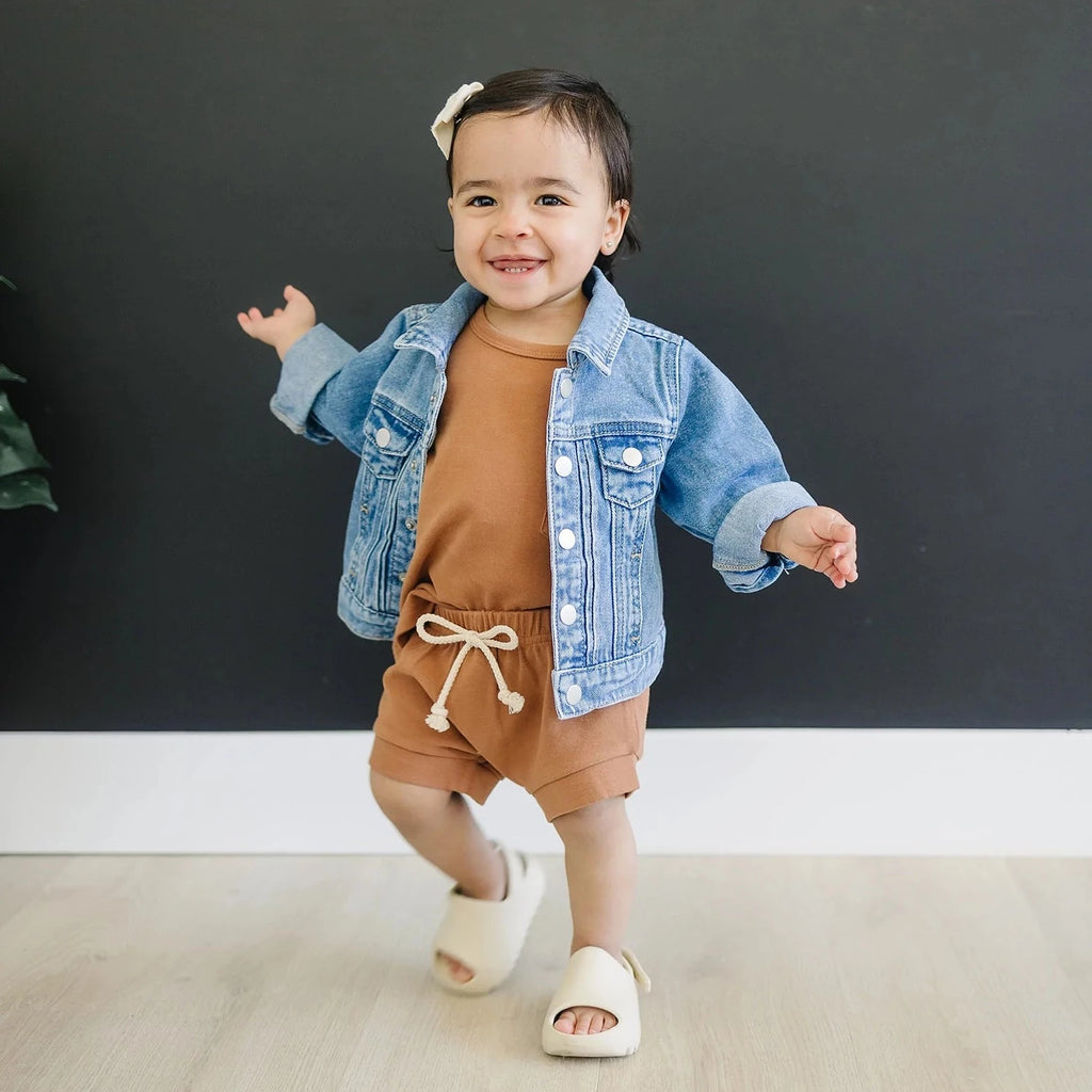 Dark background with toddler girl standing, wearing a Two Piece mebie set, with a bow in her hair, and the Mebie Baby Jean Jacket by Mebie Baby over top.