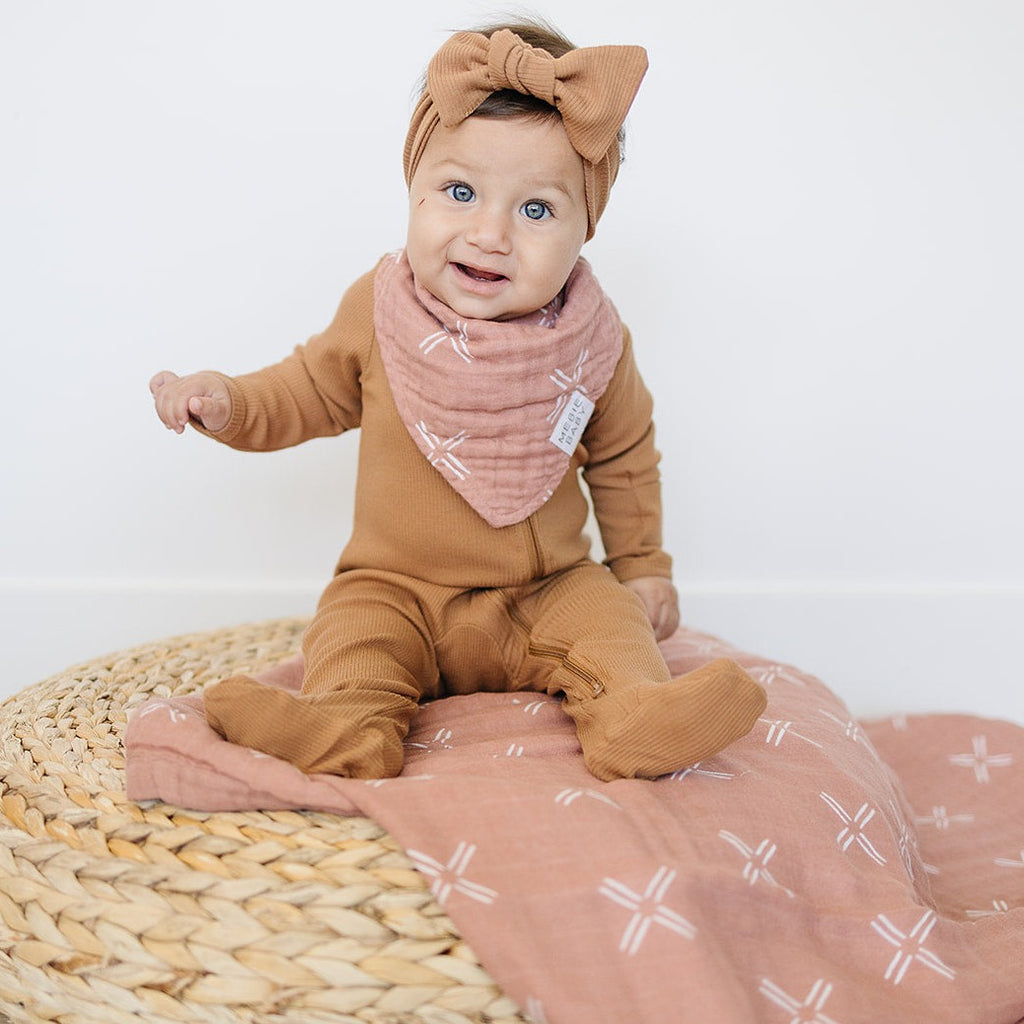 Baby wearing a Just Peachy Baby Bib by Mebie Baby, sitting on top of a wicker basket with a Just Peachy swaddle by Mebie Baby underneath. Baby is wearing a rust sleeper with a matching bow, in front of a white wall. 