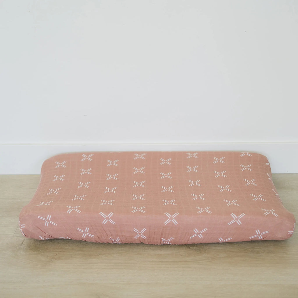 White wall with the Just Peachy Changing Pad Cover by Mebie Baby on a changing pad. Cover is a peach colour with white "x"s all over.