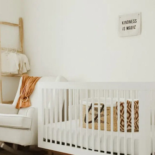 Corner of a nursery with white crib, and Kindness Is Magic Banner by Imani Collective. Banner is canvas with black text.