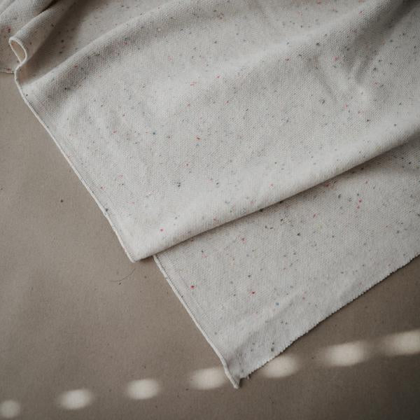 Grey background with a Knitted Confetti Baby Blanket in Ivory by Mushie. Blanket is a white with sprinkled confetti all through.