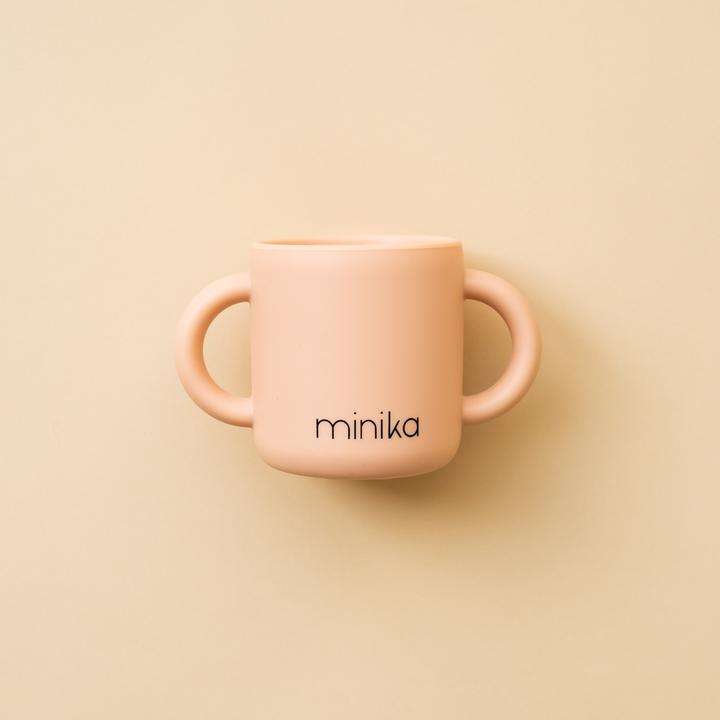 Beige background with a Learning Cup with Handles in Blush by Minika. Cup is blush silicone with 2 handles, and says “minika” in black