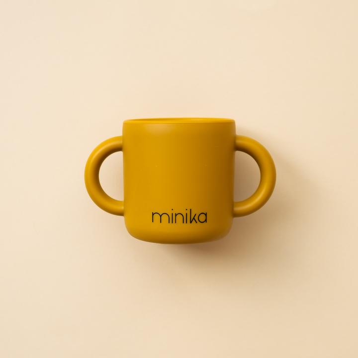 Beige background with a Learning Cup with Handles in Ochre by Minika. Cup is mustard/ochre silicone with 2 handles, and says “minika” in black.