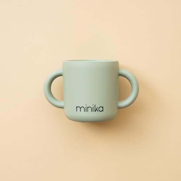 Beige background with a Learning Cup with Handles in Sage by Minika. Cup is sage silicone with 2 handles, and says “minika” in black.