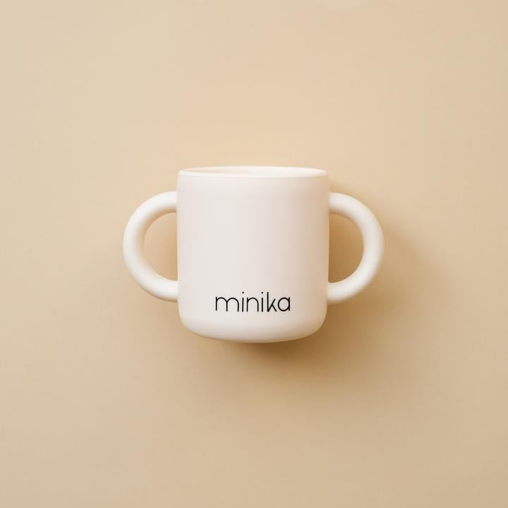 Beige background with a Learning Cup with Handles in Shell by Minika. Cup is white silicone with 2 handles, and says “minika” in black.