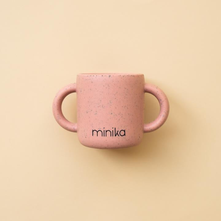 Beige background with a Learning Cup with Handles in Sorbet by Minika. Cup is pink speckled silicone with 2 handles, and says “minika” in black.