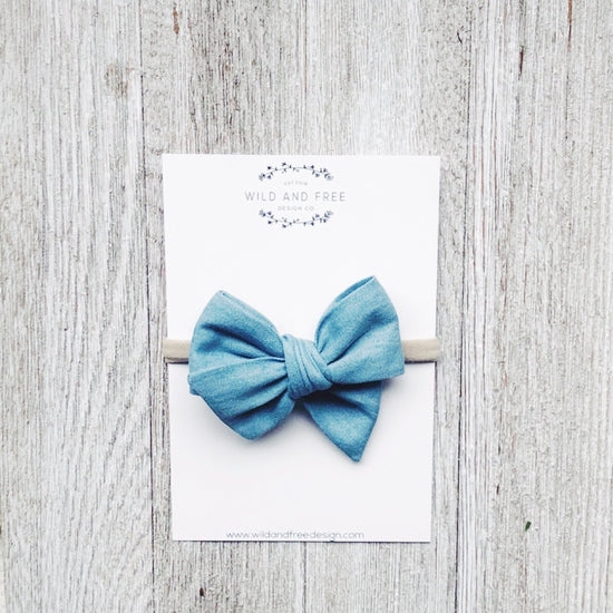 Overhead view of the Light Washed Denim Pinwheel Bow by Wild and Free Design Co. on a white washed table, in its packaging. Bow is a large pinwheel style in a medium blue denim.