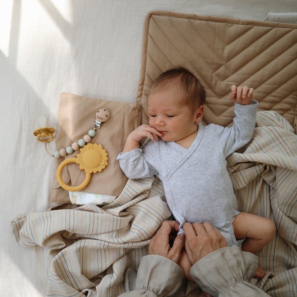 Overhead view of baby laying down on a change pad, with a swaddle, a soother and the Lion Teether by Mushie beside him.