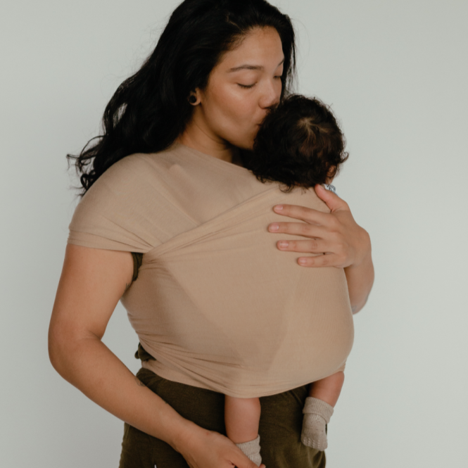 Model wearing Lennox Wrap Carrier by Beluga Baby, while holding a baby. Standing in front of a grey wall. 