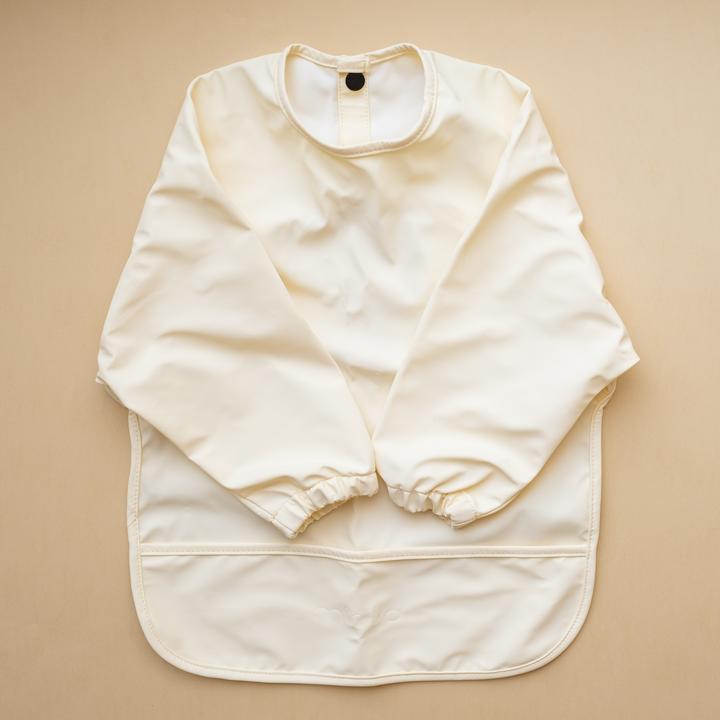 Beige background with a Long Sleeve Bib in Shell by Minika. Bib is white with long sleeves, snaps on the back, and a pouch along the bottom.