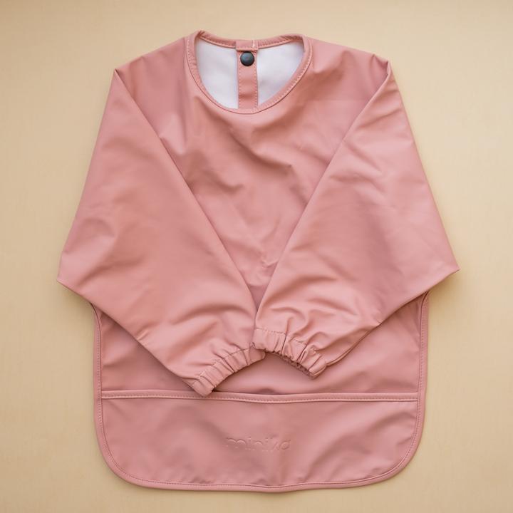 Beige background with a Long Sleeve Bib in Sorbet by Minika. Bib is rose pink with long sleeves, snaps on the back, and a pouch along the bottom.