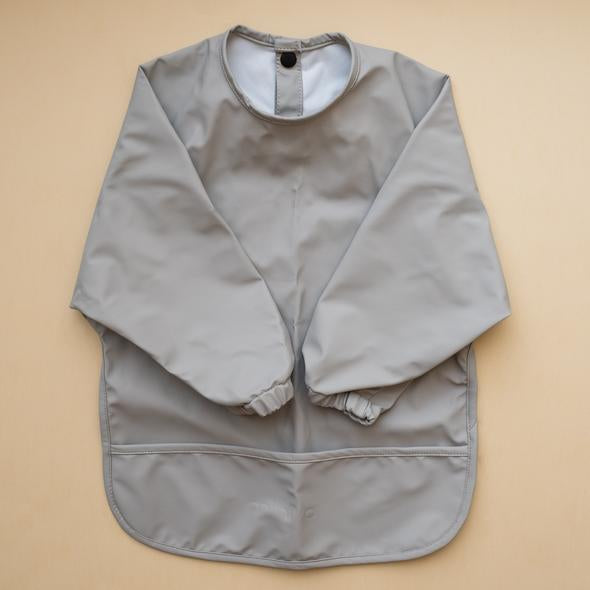 Beige background with a Long Sleeve Bib in Stone by Minika. Bib is dark grey with long sleeves, snaps on the back, and a pouch along the bottom.