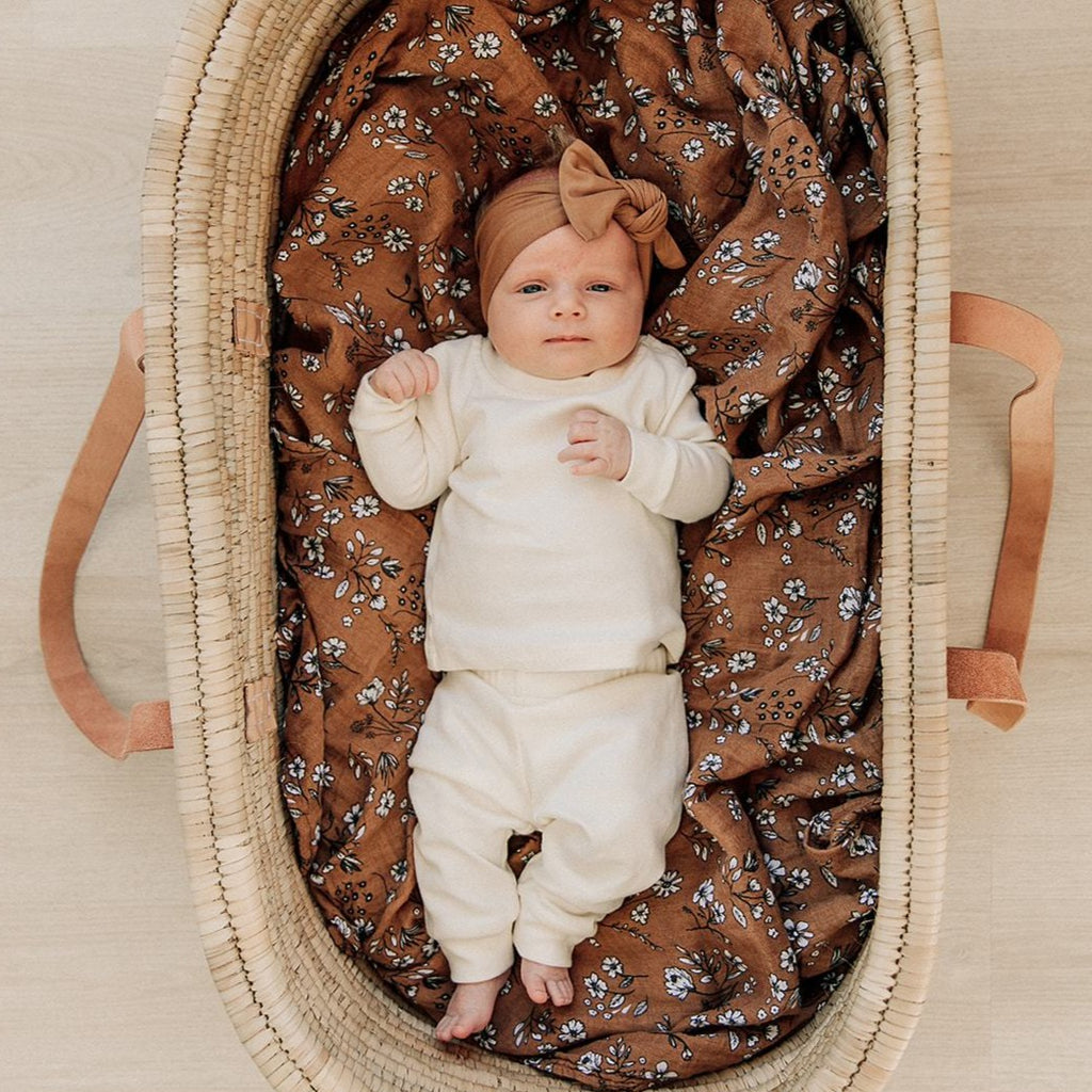 Overhead view of a baby laying in a rattan bassinet, on top of the Vintage Floral Quilt by Mebie Baby.