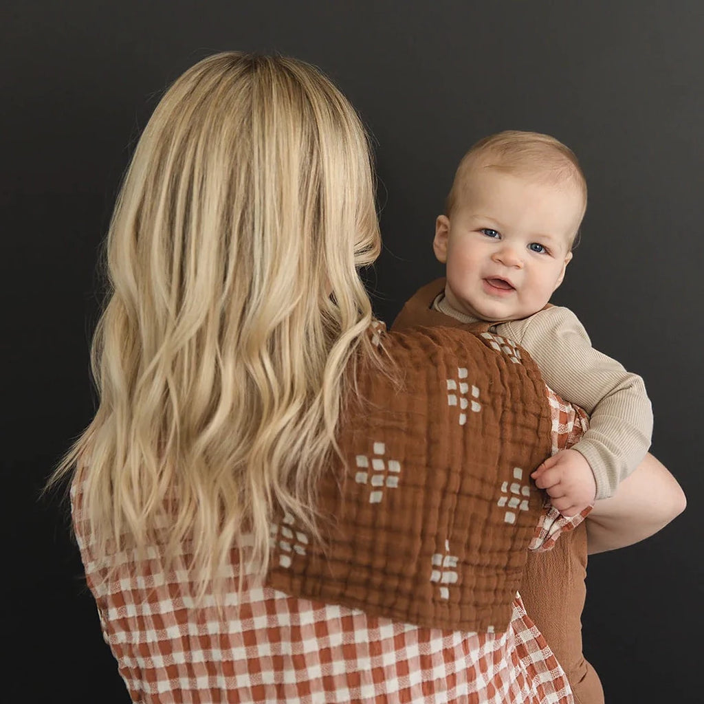Dark wall with mama, and baby standing, back to the camera with a Chestnut Textiles Burp Cloth by Mebie Baby draped over shoulder. Burp Cloth is dark brown with cream square pattern.