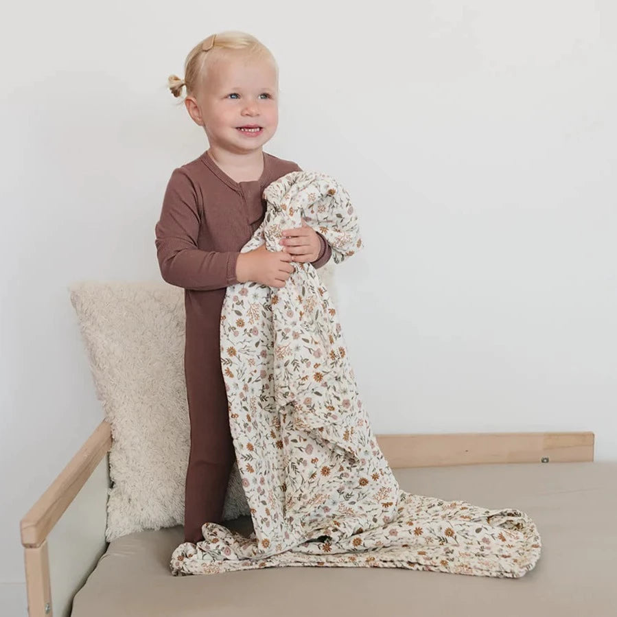 White background with a little girl standing on a natural wood bed, holding the Meadow Floral Muslin Quilt by Mebie Baby.