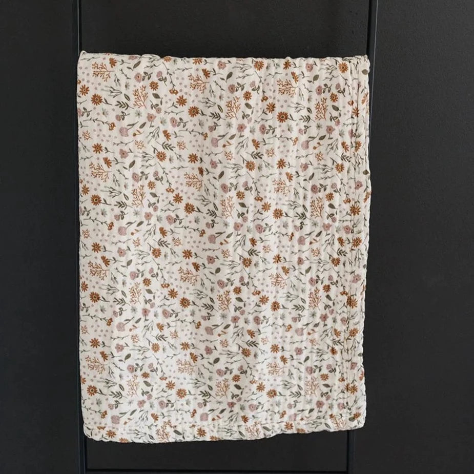 Dark background with a metal blanket ladder, and a Meadow Floral Muslin Quilt by Mebie Baby hanging on it. This quilt is a cream colour with rust, green, and mauve flowers.