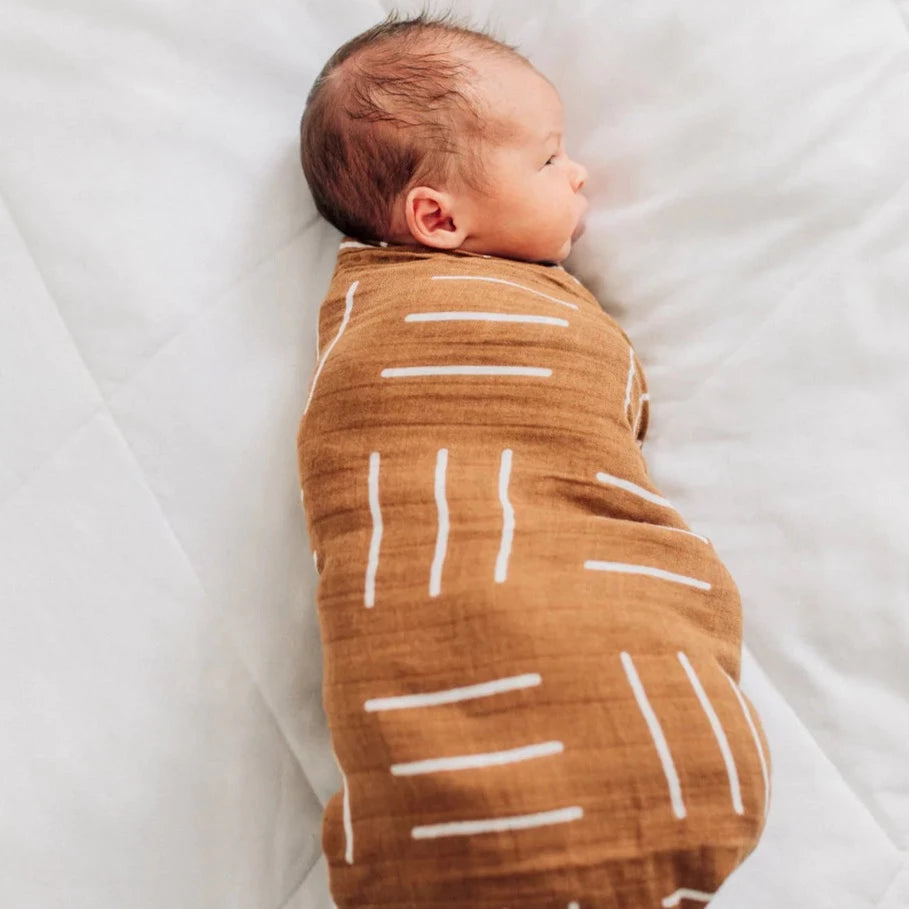 White background with overhead view of baby wrapped in a Mustard Mudcloth Muslin Swaddle by Mebie Baby. Swaddle is mustard with white lines.
