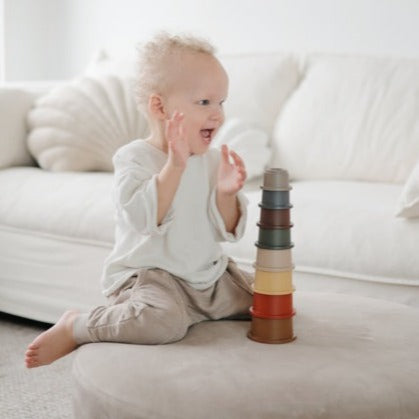 Little boy sitting on a white couch & playing with the Stacking Cups Toy by Mushie.
