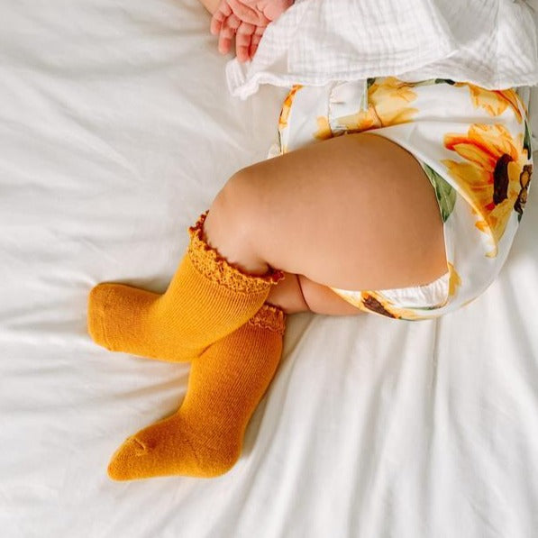 White blanket background with close up of baby laying on her side, wearing a sunflower romper with a pair of Lace Knee High Socks in Mustard by Little Stocking Co.