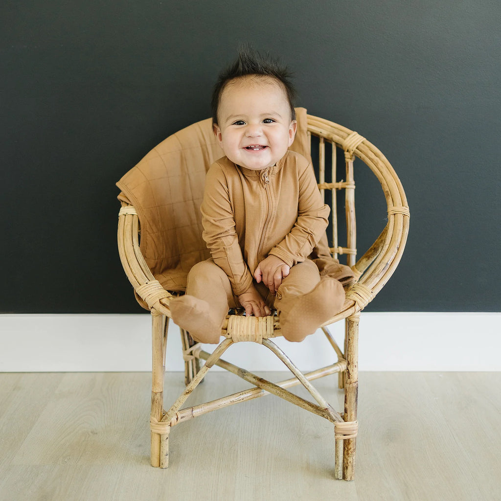 Dark wall with a rattan egg chair in the middle, and a baby boy sitting in it with the Mustard Bamboo Quilt by Mebie Baby laying over the back.