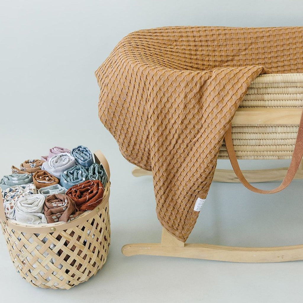 White background with a rattan bassinet and a Mustard Waffle Quilt by Mebie baby laying over it.