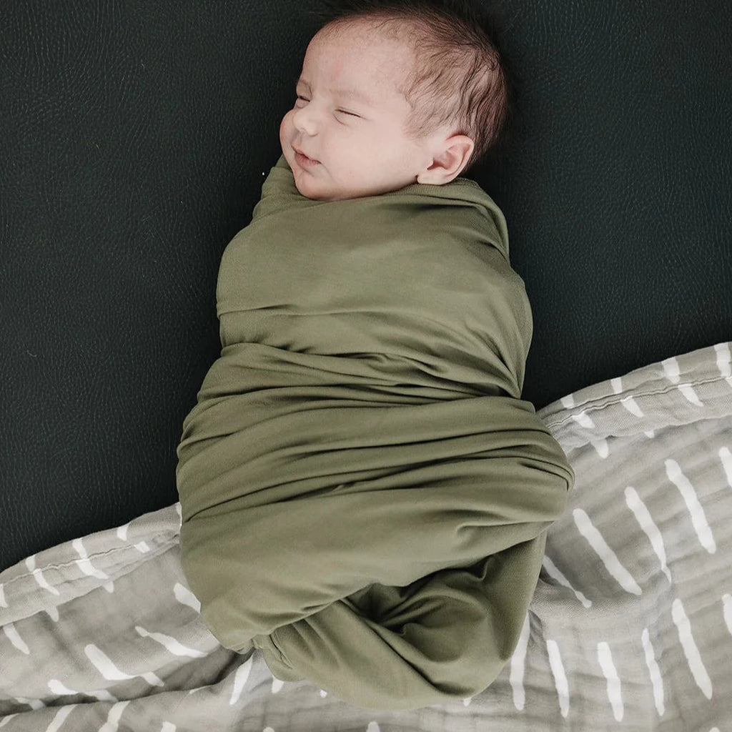 Overhead view of baby laying on black leather, wrapped up in an Olive Stretch Swaddle by Mebie Baby. Swaddle is olive, and has lots of stretch.