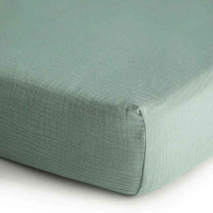 White background with a close up of the corner of a mattress with the Extra Soft Muslin Crib Sheet in Roman Green by Mushie on it. Roman Green is a eucalyptus green.