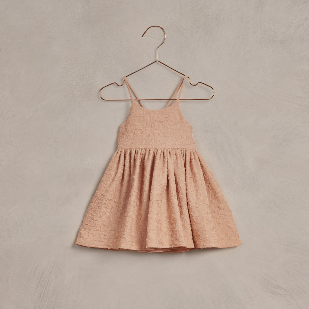 Pippa Dress in Blush by Noralee, hanging on a rose gold hanger on a grey wall. 
