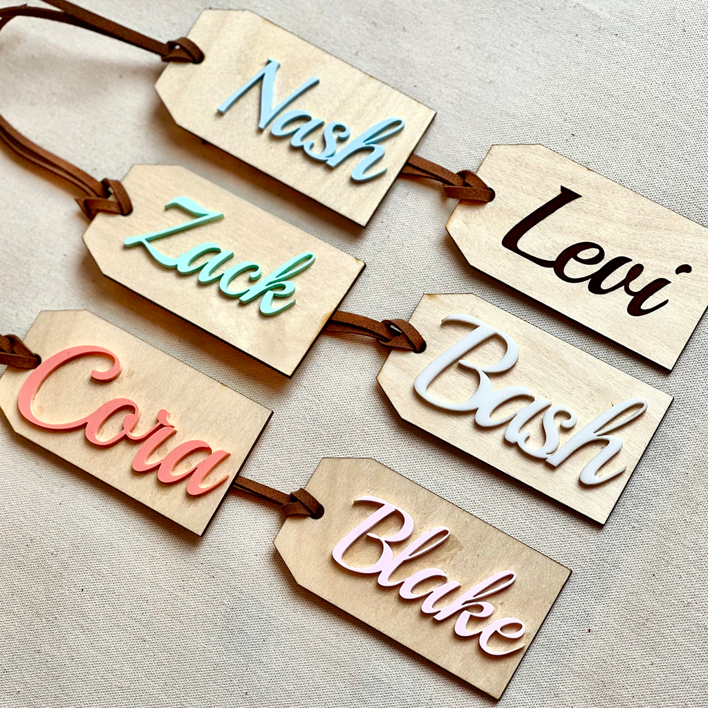 Customized Name Tags by Petit Nordique in Birch Wood with vegan leather. Laid on a beige surface. Names from left top to bottom. Nash in Sky, Levi Engraved, Zack in Seafoam, Bash in Cloud, Cora in Coral and Blake in Rosette. 