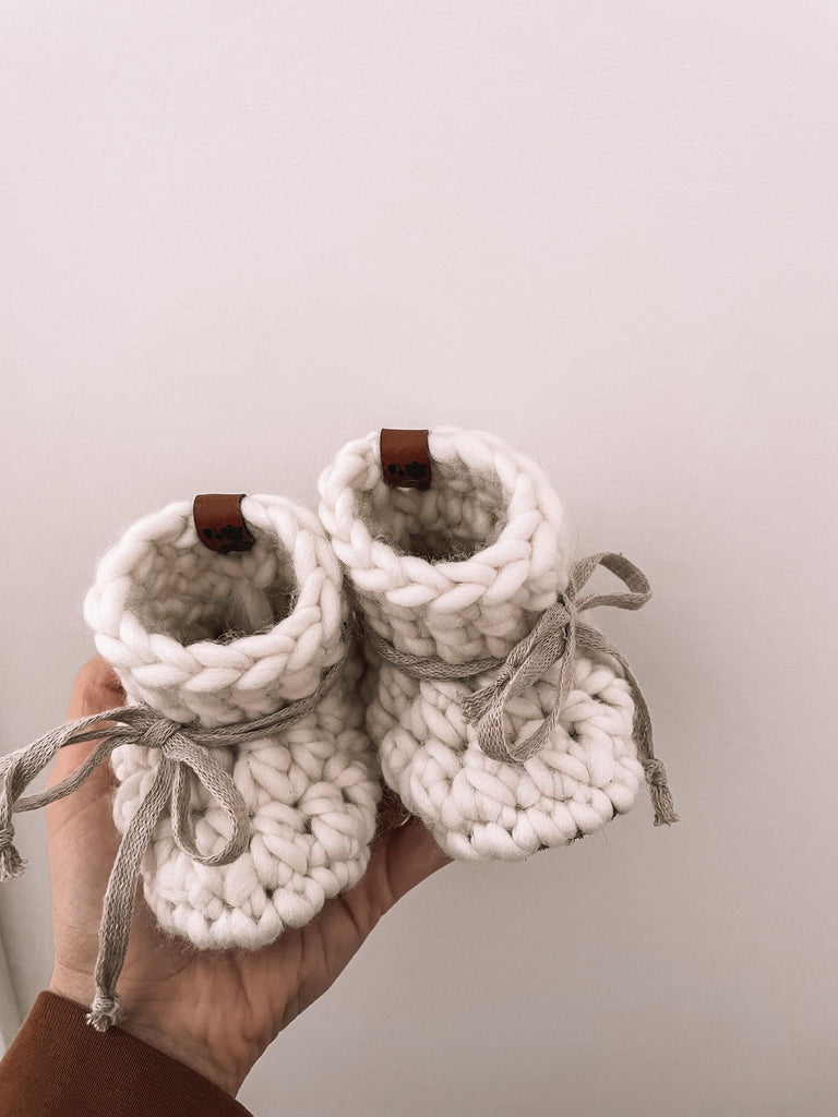 Natural Baby Booties by Petit Nordique with greige ties and brown leather tags, being held up by a hand against a white wall. 