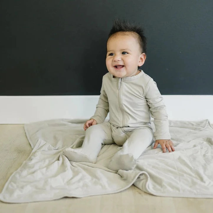 Dark wall with a light wood floor, and a baby sitting on a quilt wearing the Oatmeal Bamboo Zipper One-Piece by Mebie Baby.