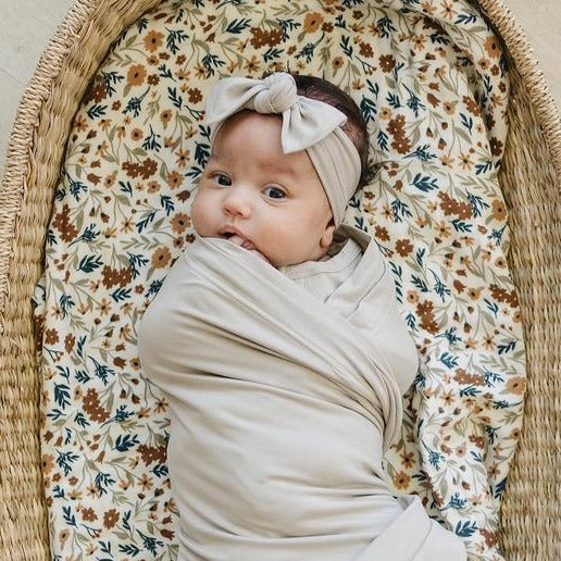 Overhead view of baby laying in rattan bassinet, wearing an Oatmeal Head Wrap by Mebie Baby. Head wrap is wide, with a knot bow at the front in a beige/grey colour.