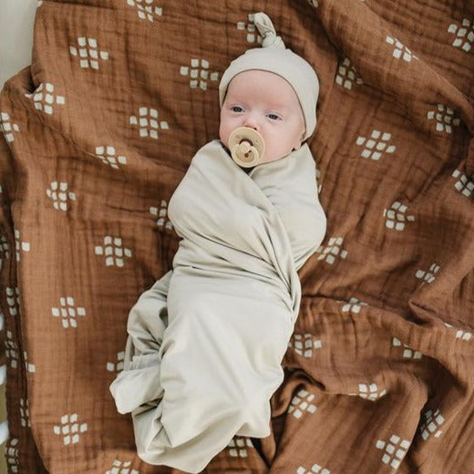 Overhead view of baby laying down wearing an oatmeal Newborn Knot Hat by Mebie Baby. Knot hat is a pale grey/beige, and fits snug with a knot at the top.