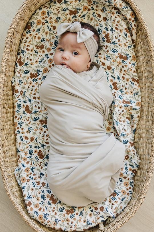 Overhead view of baby laying in bassinet, wrapped up in Oatmeal Stretch Swaddle by Mebie Baby. Swaddle is a pale grey/beige colour, and is full of stretch.