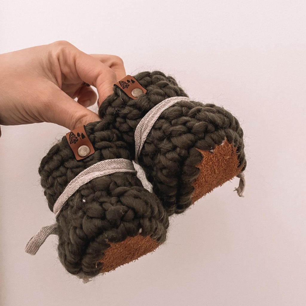 Olive booties with leather soles by Petit Nordique. Booties have leather tags and greige ties, being held up by a hand in front of a white wall.