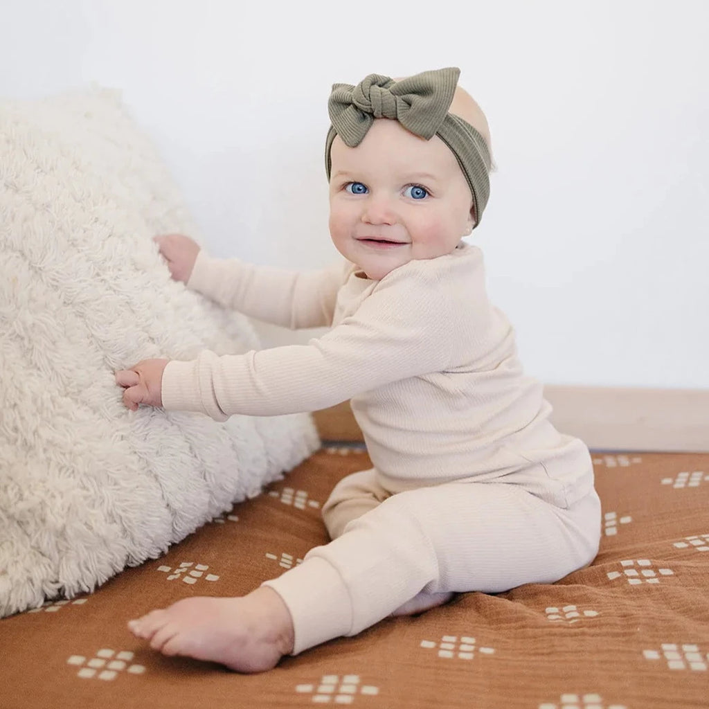 White background with a baby girl sitting, wearing the Olive Head Wrap by Mebie Baby.