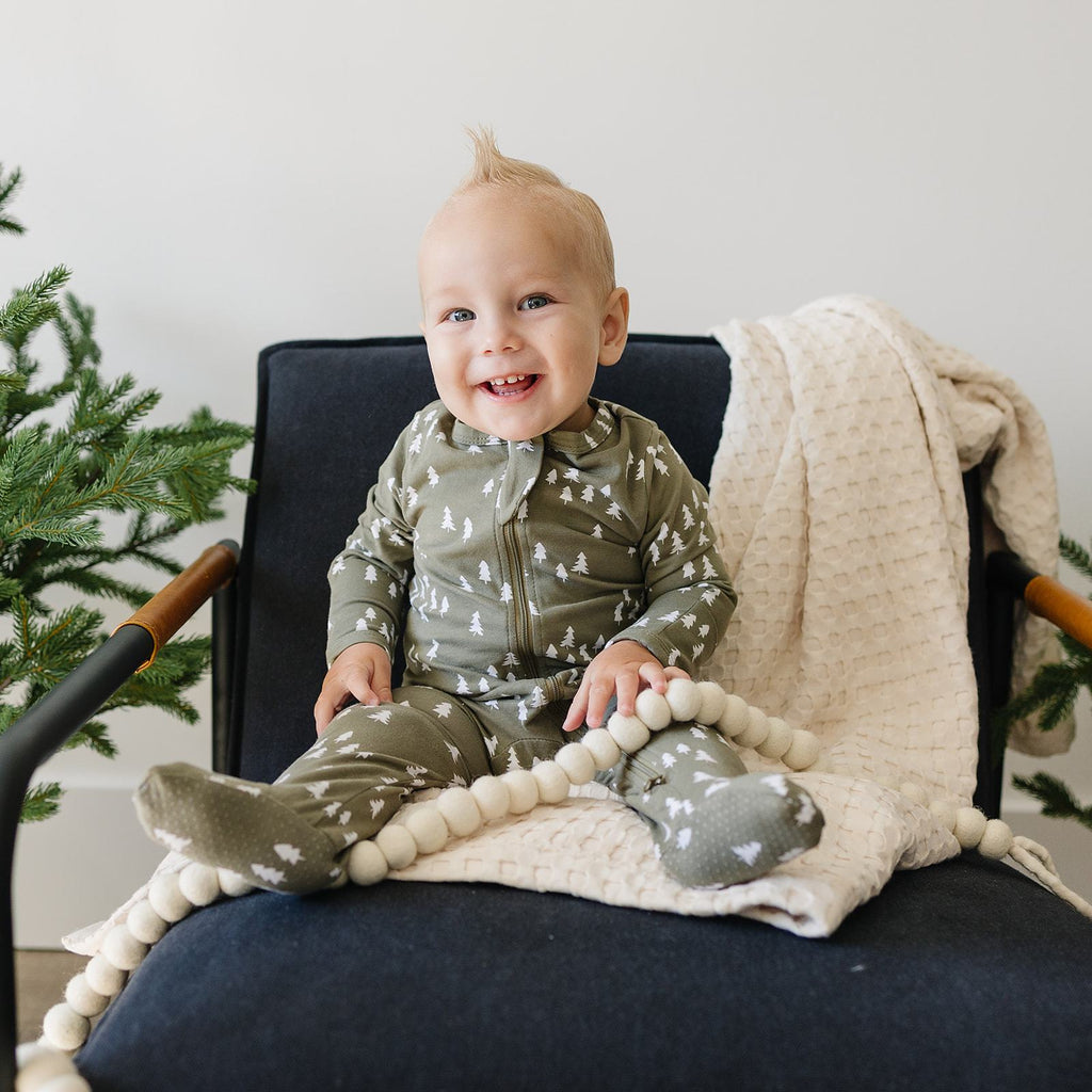 White wall with a navy blue chair, and a little boy sitting on it smiling, wearing the Olive Pines Footed Zipper One-Piece by Mebie Baby