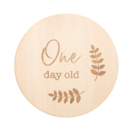 Round wooden milestone disc that says "One Day Old" with sprigs of greenery by Bebe Au Lait