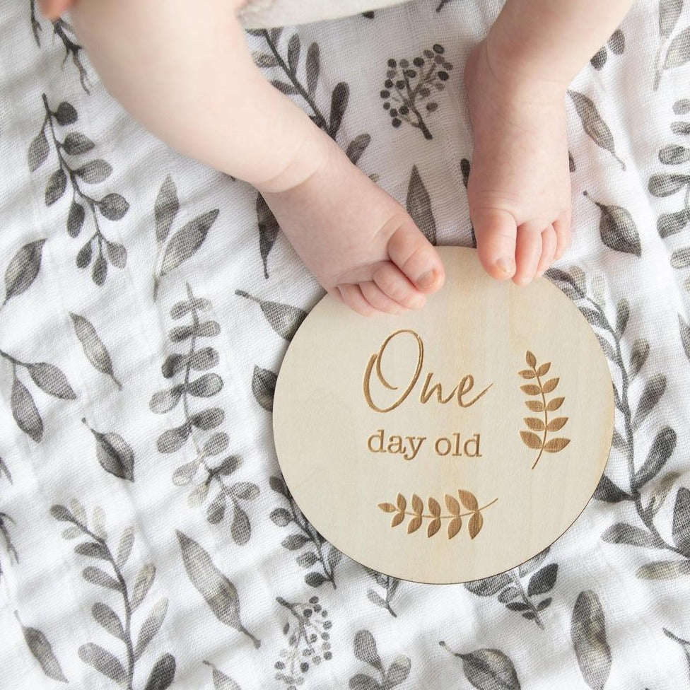 Baby toes on a blanket with round wood milestone disc that says "One Day Old" with sprigs of greenery by Bebe Au Lait