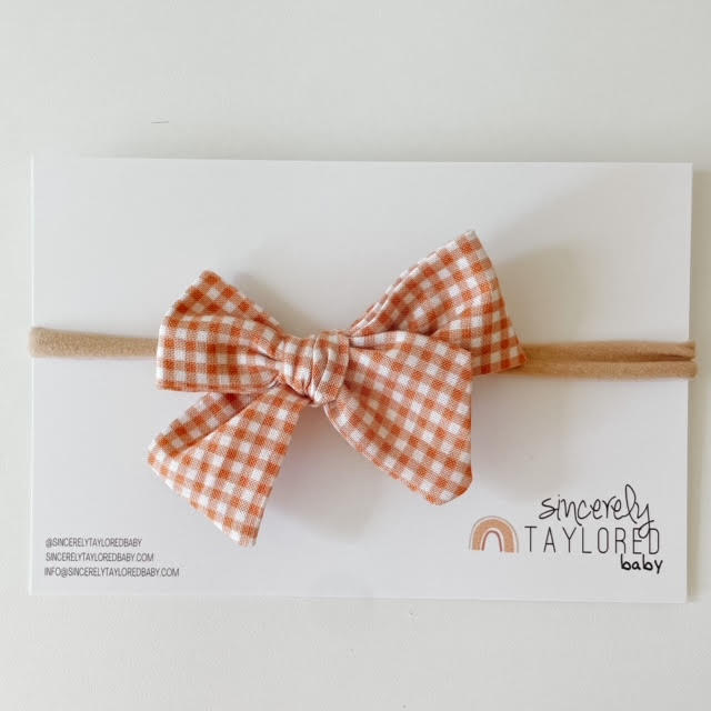 White background with the Orange Plaid Bow by Sincerely Taylored Baby, in its packaging. Bow is an orange and white plaid, on a nylon headband.